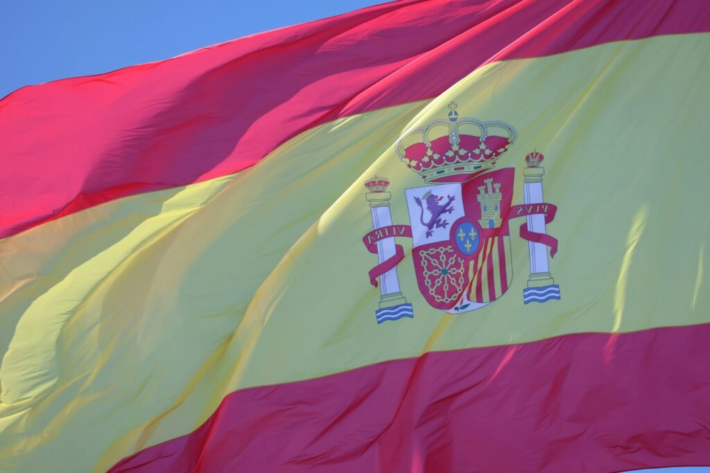 A vibrant Spanish flag with coat of arms waving against a clear blue sky in Cádiz, Andalucía.